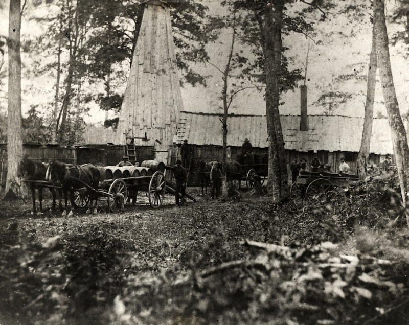 Two teams of horses in front of carts loaded with wooden barrels. A group of men is loading the barrels onto the carts. There is a large building in the background