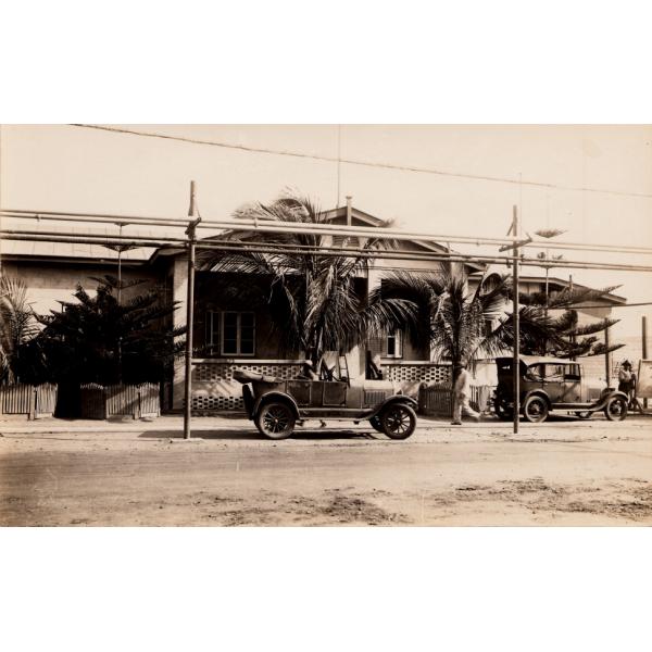 A photo of a single-story buildings with latticework on the porch and palm trees in front. There are two cars parked outside beside a dirt road. Hydro wires run overhead. 