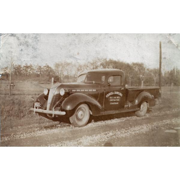 A two-door truck parked on the side of a dirt road with a field and trees in behind it. The truck is dark and has "Stover and Rawlings. Oil & Gas Well Contractors. Chatham" written on the door. 