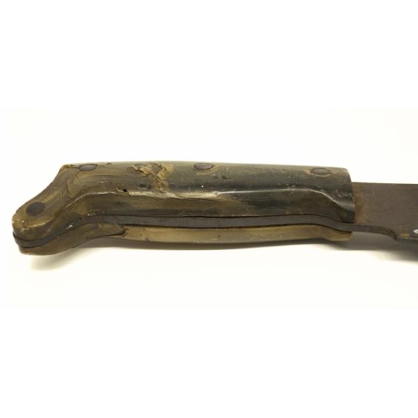Vue de près du manche en os de la machette. Il y a un morceau d’os des deux côtés de la lame de métal, et des morceaux métal fixent l’os sur le métal.