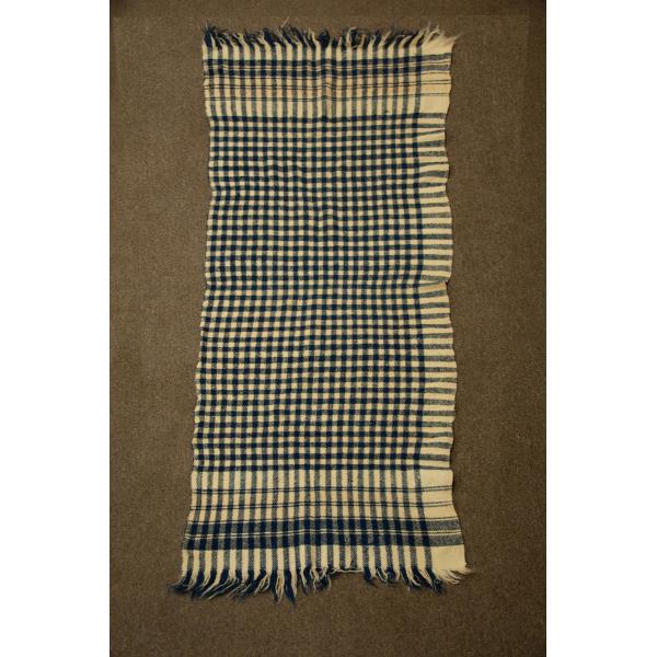 Une couverture quadrillée beige et marine. Les bordures du haut et du bas sont ornées d’une frange. 