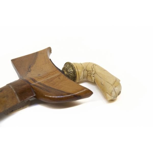 The carved ivory handle of a dagger. There are lines on it and it is curved. There is a brass-coloured metal piece with swirling lines connecting it to the dagger. The brown wooden sheath is shaped like a "T". 