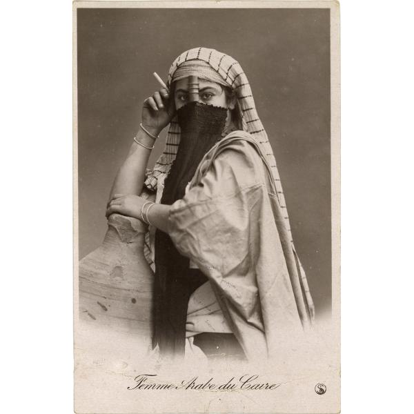 A postcard with a veiled woman wearing a white striped cloth on her head. She has two bracelets on each wrist and is holding a cigarette. The bottom reads: "Femme Arabe du Caire".  