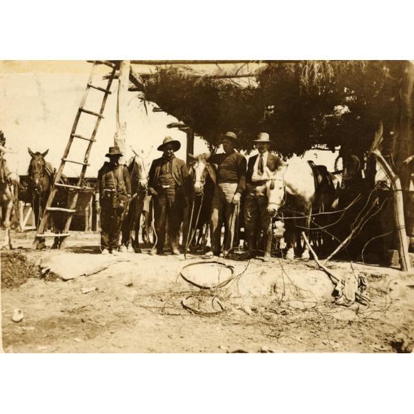 A photo of four men who are wearing hats standing with five horses, underneath a structure with a roof made from vegetation that has a wooden ladder leaning against it.