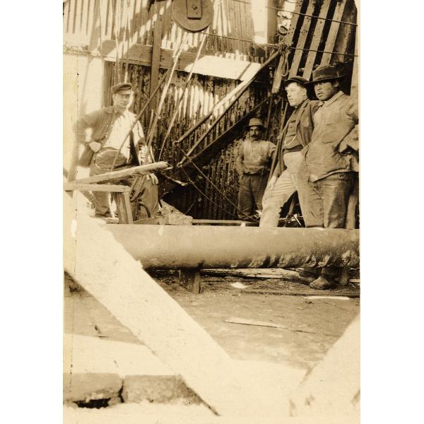 Four men stand inside of an oil rig that is covered with sheet metal. There is a large pipe running horizontally a foot from the floor.