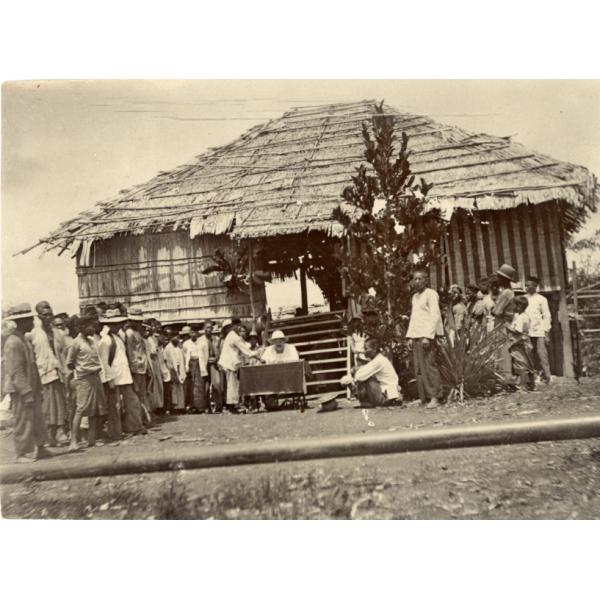 Group of local labourers living up to the left to receive payment from William McRae who is sitting at a desk in front of a hut.