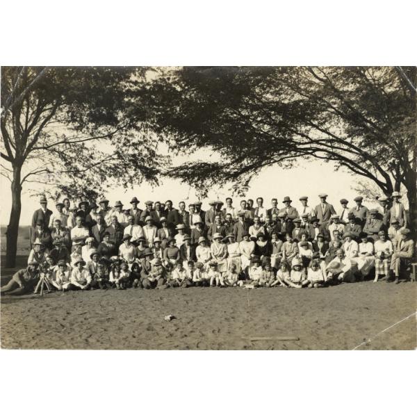 Group picnic at the Chira River. There is sand in front of them and two trees in behind. George Rawlings is standing in the back row, third from the left, in a pith helmet. 