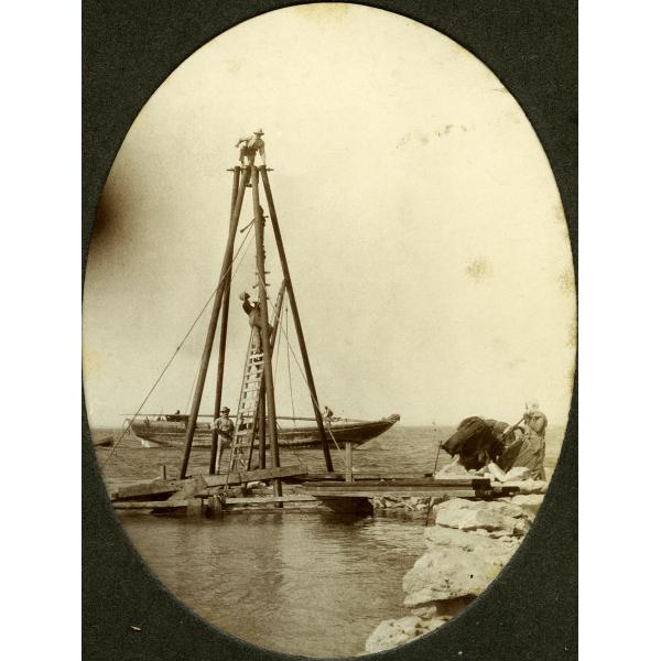 A photo of three men on a wooden tower wit four poles and ropes leading down to a dock. A boat with its mast down is anchored in the water behind them. There is a fourth man on the right beside a pile of objects wrapped in dark material.