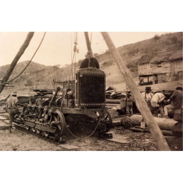 A photo of a partially assembled bulldozer beneath a wooden tripod. There is one man at the front of the bulldozer and four behind it. There are hills in the background. 