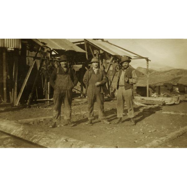 Three International Drillers wearing work clothes and hats, standing in front of an oil derrick with a roof made from metal sheeting. There are dirt hills in the background.