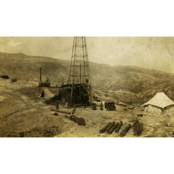 A metal oil rig. There are men standing around it and near a car. There are metal cylinders in the foreground and a tent on either side of the rig. There is a cylindrical steam boiler behind the rig. There are hills in the background.