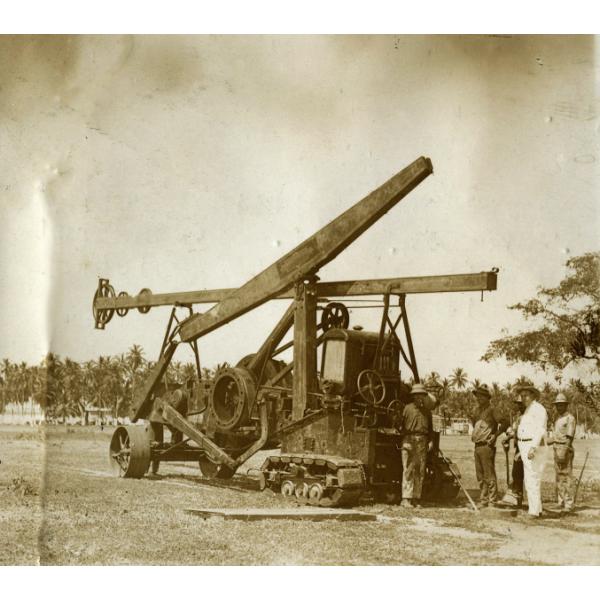 A photo of a star drilling machine with a tractor in front. There are five men standing on the right-hand of the photo and there are trees in the background.