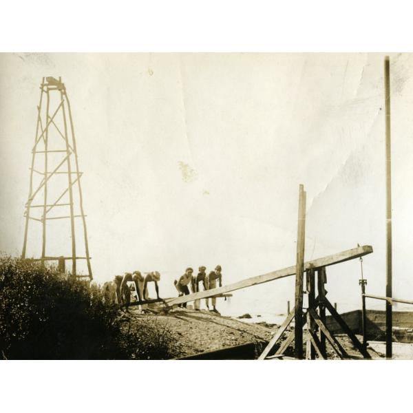 A photo of six men pushing down on a wooden crossbar supported by a wooden frame. There is a wooden four-pole structure behind them and a bush to the right.