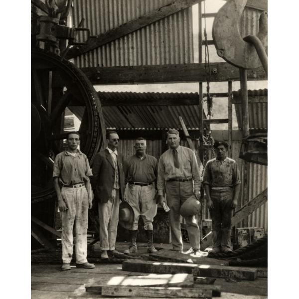 Group of five men in dirty clothing standing inside an oil rig covered in sheeting. There is a large wheel and belt behind them. 