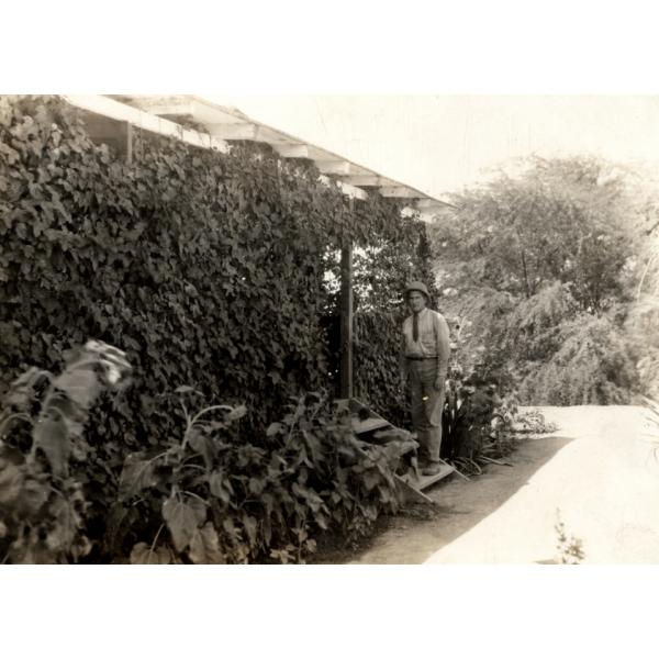 A photo of George Rawlings in a suit and pith helmet standing on the wooden steps of a vine-covered house. 