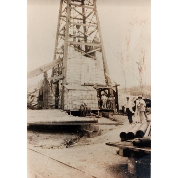 A photo of the bottom half of an oil rig being thatched. A group of men stand at the base. There are piles of casing on either side. 