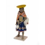 Front view of a figurine of a woman wearing sandals and who is dressed in a blue skirt with an embroidered design along the edge, as well as an orange rectangular shoulder cloth fastened at the front. She has an inverted yellow hat and is carrying a spool and wool. 
