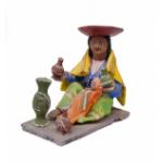 A figurine depicting a woman wearing a green skirt, a purple blouse, a blue vest with a design on it, and a yellow rectangular shoulder cloth. She is wearing an inverted red hat. There is a baby in her lap and she is holding pieces of pottery. 