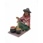 Figurine of a woman stirring two black pots with a brown spoon. She is wearing sandals, a green skirt, and a red rectangular shoulder cloth. There is a white apron wrapped around her wrist and she is wearing a grey hat with the brim curled up. 