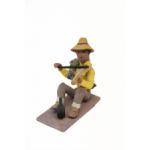 Figurine of a man playing the fiddle wearing sandals, brown cuffed pants, a yellow shirt, and a green bag tied over one shoulder. He has a yellow hat and there is a black bottle by his feet. 