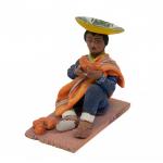 Figurine of a man eating fruit wearing sandals, blue pantsuit, a yellow sash, with an orange poncho thrown over one shoulder. He has an inverted yellow hat and there are four orange egg-shaped objects by his foot.