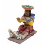 Figurine of a woman eating with two chickens in front of her. She is wearing a red skirt, yellow shirt, and blue rectangular cloth around her shoulders. She has an inverted orange hat on. 