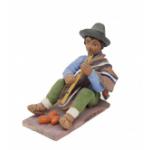 Figurine of a man playing the flute wearing sandals, green pants, a blue shirt, and a brown poncho over one shoulder. He has a grey hat and there are three orange egg-shaped objects at his feet. 