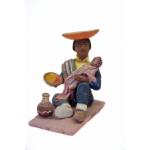 A figurine showing a man sitting on a pink mat and feeding a baby. He has an inverted orange hat and is wearing sandals, blue pants, blue shirt, yellow vest, and green poncho thrown over one shoulder. There is a vase by his right foot.