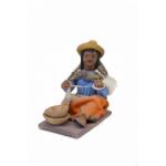Figurine of a woman spinning wool wearing sandals, an orange skirt, a white apron, a blue blouse, and a brown rectangular shoulder cloth. She has a yellow hat and there are two brown bowls by her feet.  