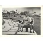 A photo of Vola and John Braybrook sitting on a park bench. There is a path with large stones beside them and a series of benches along the path behind them. She is wearing a skirt and jacket with high heels and he is wearing a suit and tie.
