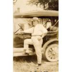 A photo of Fred Webb standing with his foot up on the step of a car. Webb is wearing a white shirt and pants, a tie, and a boat hat. The car has a canvas top and no windows. There is a building with a peaked roof in the background. 
