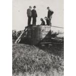 A group of five men standing on an above-ground wooden storage tank. There are metal rings around the barrel holding it together. There is a ladder propped against the tank so the men could climb on top of it.