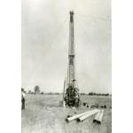 A photo of a small drilling rig in a field. There are four wires extending from the top of the rig down to the ground. Three men are looking at the rig and there are pieces of casing laying on the ground in front of it.
