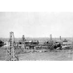 Three oil derricks stand in a field in front of a group of buildings. There are pipes connecting the buildings. At the bottom of the picture, "Woodward's Refinery" is written in white.