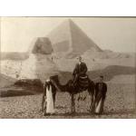 John Growder sitting on a camel in front of the Sphinx and a pyramid. There is a man holding the reins of the camel and another man at the back of the camel.
