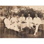Cinq hommes sont assis autour d'une table en osier recouverte d'une nappe blanche. Un bouquet de fleurs est posé devant un cadre sur la table. Les hommes portent des costumes blancs.