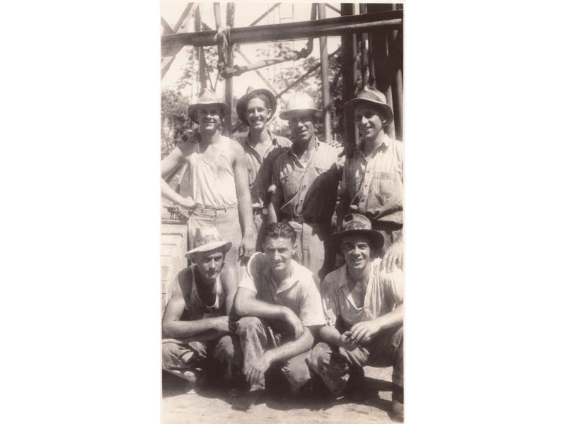A group of seven International Drillers underneath a metal frame. Four are standing and three are kneeling. They are wearing work clothes.