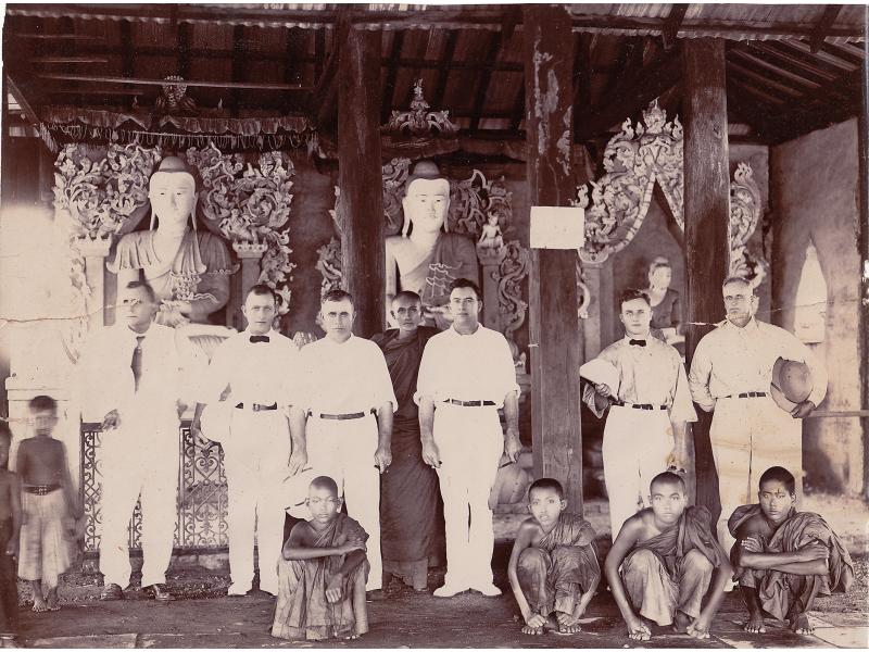 Six foreurs internationaux vêtus de blanc à l’intérieur d’un temple, avec cinq jeunes garçons et un moine. Il y a trois piliers allant du sol au plafond et trois statues en arrière-plan.