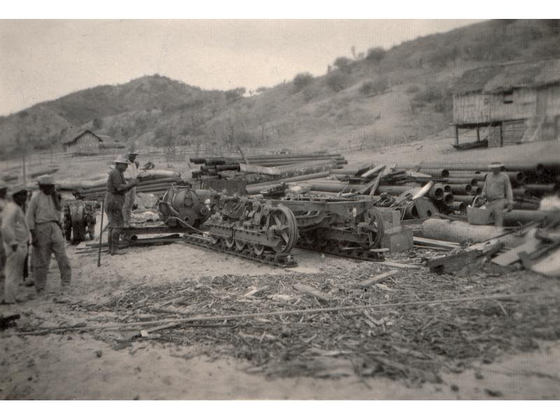 A photo of a half-assembled bulldozer. George Rawlings stands to the left. There are stacks of pipes in behind the bulldozer and two buildings on the hill at the back of the scene.