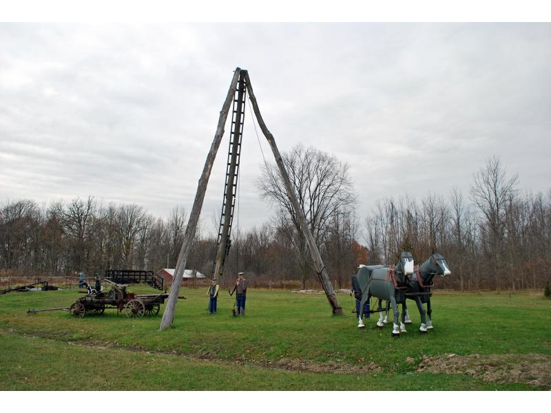 A three-pole derrick with tin figures around it, showing a team of horses, man at the pump head, and a boy and dog sitting on a portable pump kit.