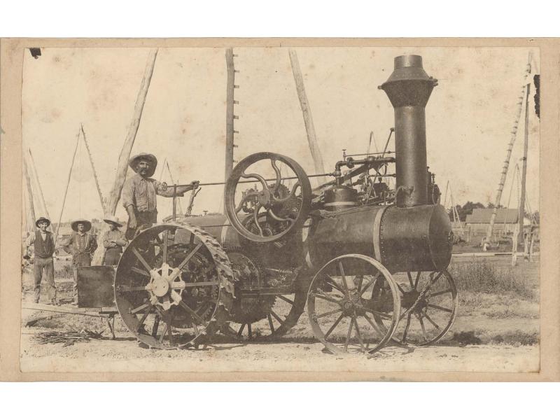 Four men standing at the back of a boiler. It has larger wheels in the back and smaller wheels in the front. There is a chimney at the front and a large gear in the middle. There are three-poled derricks in the background.