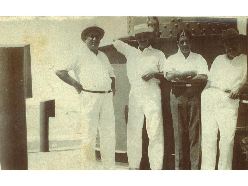  A photo of four men dressed in white on the deck of a ship. They are standing in front of a metal column with rivets in it.