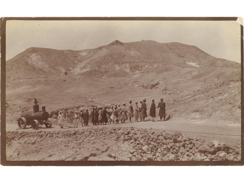 A group of men pulling a boiler with a rope up a slight incline in the desert. There are stones on the road and hills in the background.