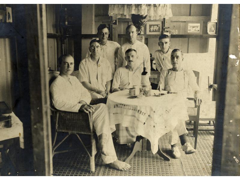 A photo of seven International Drillers around a table indoors with a white tablecloth. They are wearing white and four are sitting on wicker chairs. There is a cup, a pipe, and a bottle on the table.