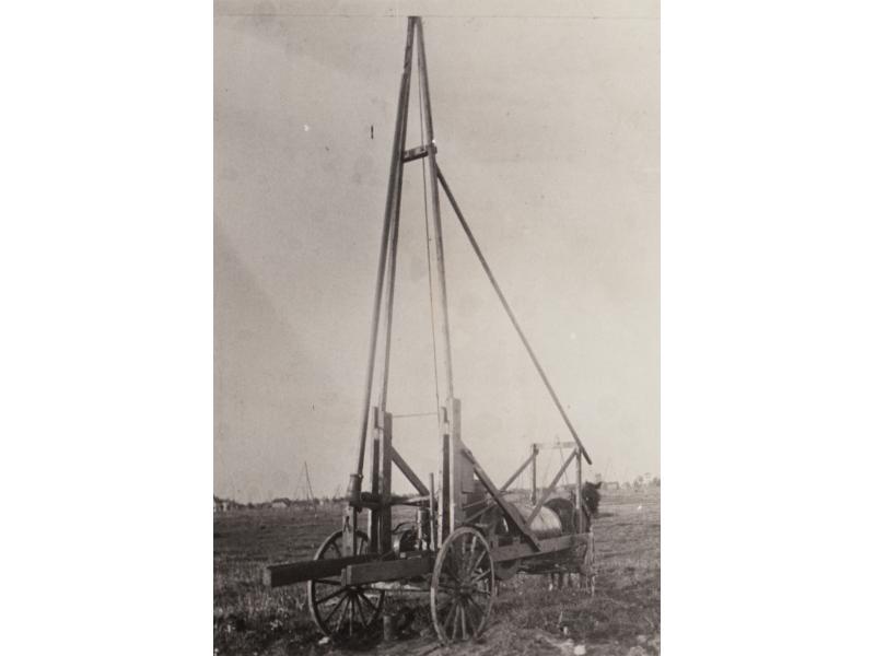 A crude oil dipping machine on a cart with four wheels being pulled by a horse. There are three main poles and a support pole running down towards the horse.