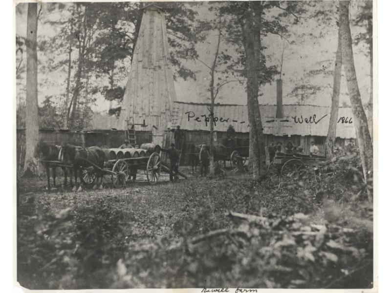 Two teams of horses stand in front of carts loaded with wooden barrels as men add more barrels. There is a large building behind them and "Pepper Well 1866" is written across the photo.