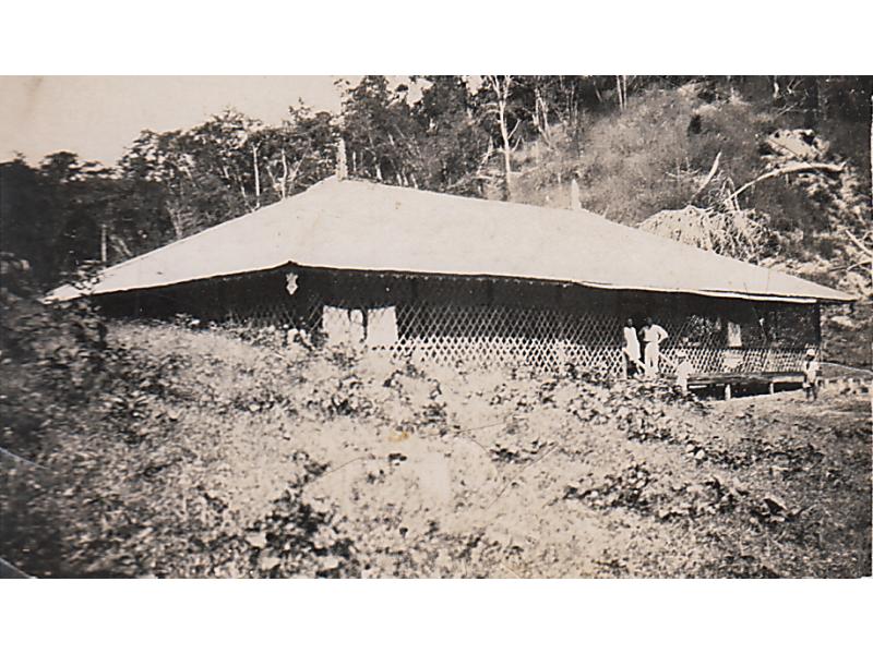 La maison de plain-pied est sur une colline, avec un toit léger et un treillis l’entourant. Trois personnes se tiennent debout dans les escaliers et une autre à quelques mètres.