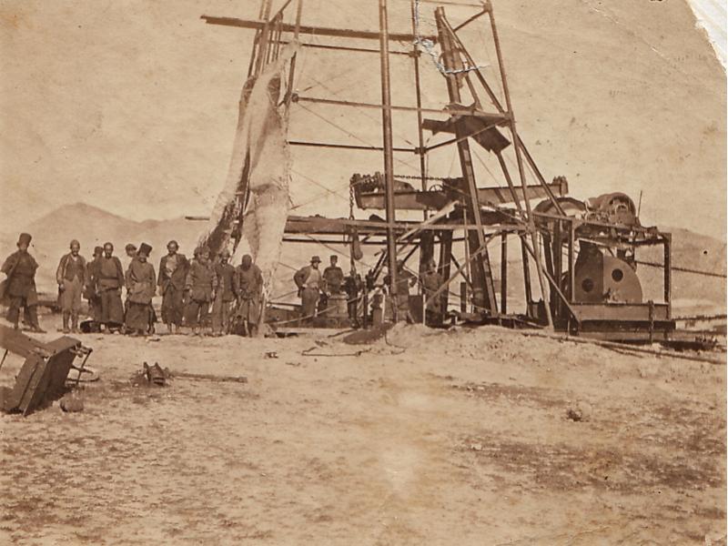 Une photo d’un groupe d’homme portant des habits de travail et se tenant à la base d’une installation de forage pétrolier en métal. Une étoffe recouvre le côté droit de l’installation de forage. Le sol est nu et il y a des collines en arrière-plan.
