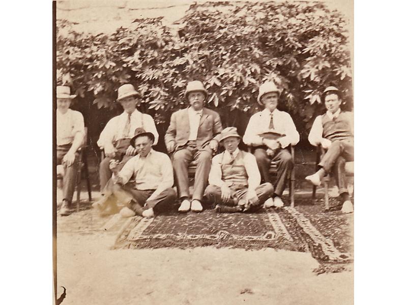 Une photo de trois tapis à motif sur le sol. Cinq foreurs internationaux sont assis sur des chaises et deux autres sont assis sur le sol. Il y a des arbustes derrière eux.