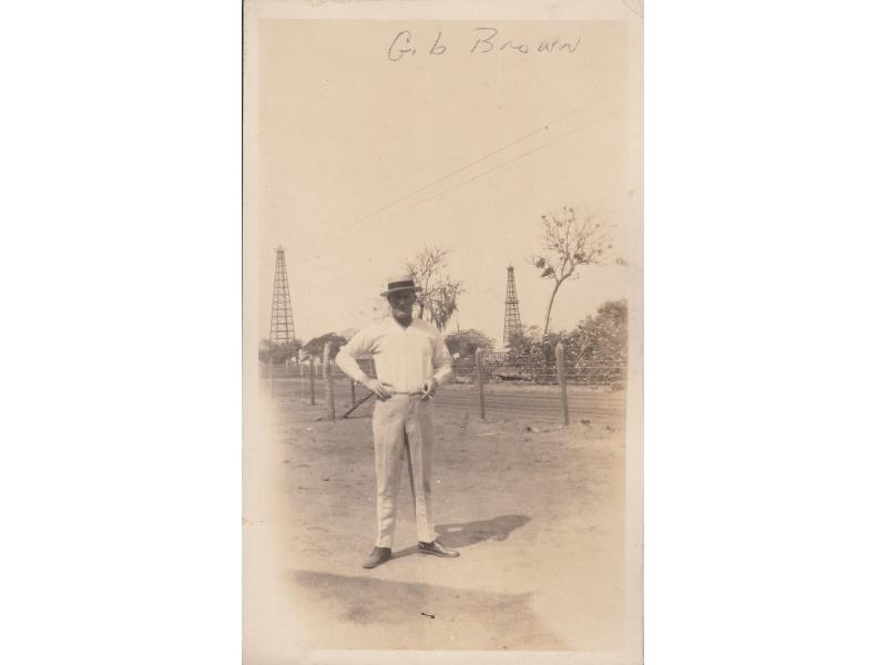 Gilbert Brown standing in front of a wire fence, holding a cigarette. He is wearing a white shirt and pants, with a boat hat. There are two oil derricks in the background.